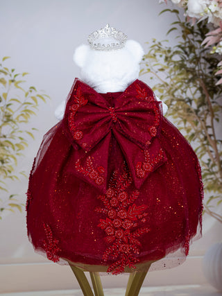 A burgundy dress displayed on a white teddy bear, featuring a large red bow at the back, glittery tulle skirt, floral embroidery, and a small silver tiara on top. The bear is set against a soft, light background with decorative plants.