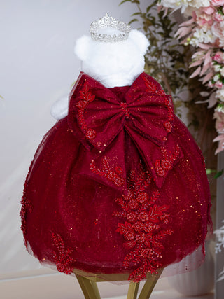 A burgundy dress displayed on a white teddy bear, featuring a large red bow at the back, glittery tulle skirt, floral embroidery, and a small silver tiara on top. The bear is set against a soft, light background with decorative plants.