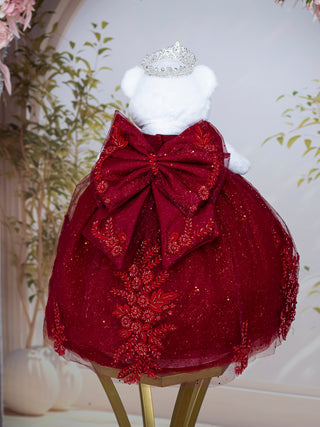 A burgundy dress displayed on a white teddy bear, featuring a large red bow at the back, glittery tulle skirt, floral embroidery, and a small silver tiara on top. The bear is set against a soft, light background with decorative plants.