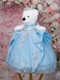 White teddy bear in a blue dress with lace details and a tiara, standing against a neutral background.